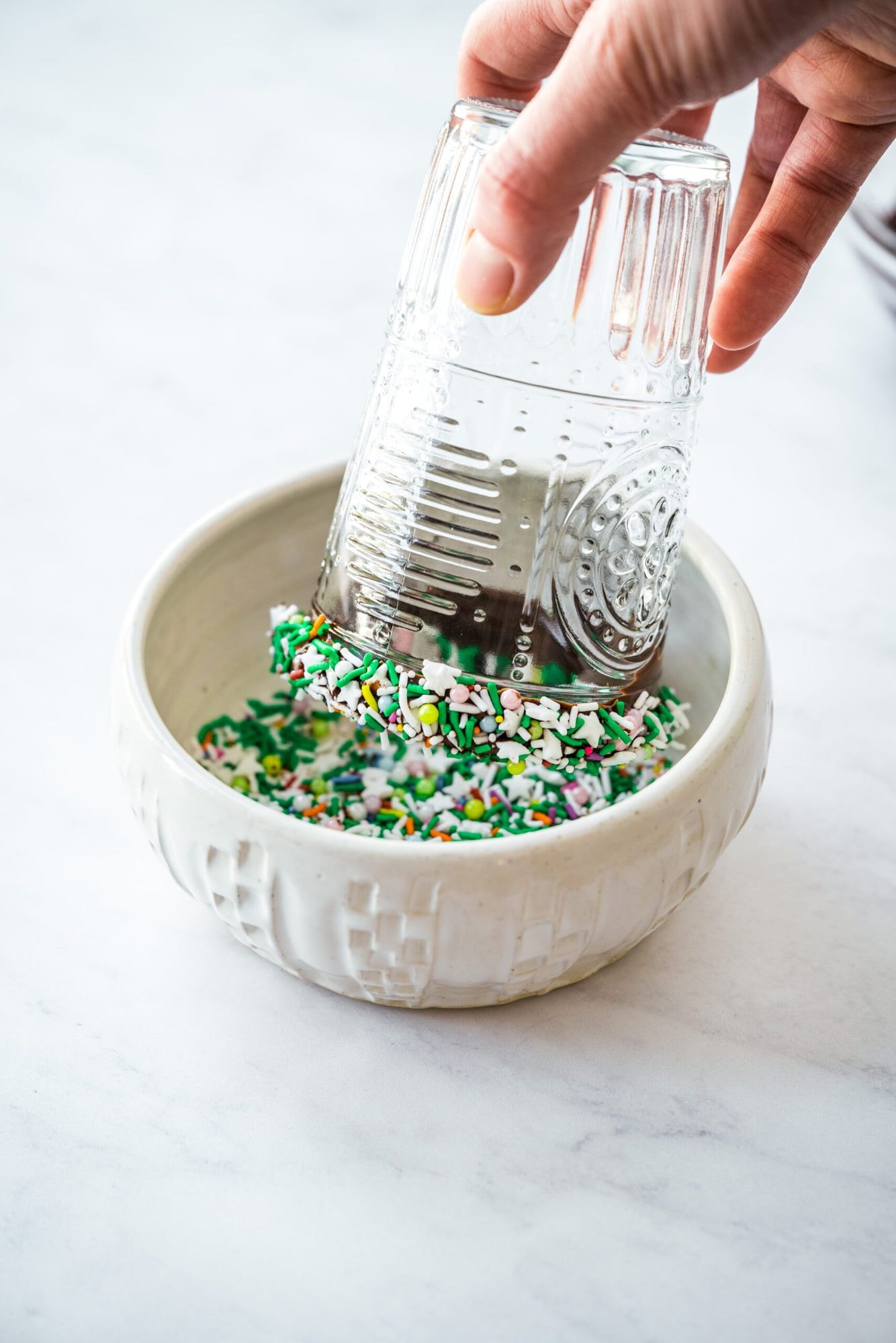 person dipping chocolate-coated rim of glass into bowl of sprinkles. 