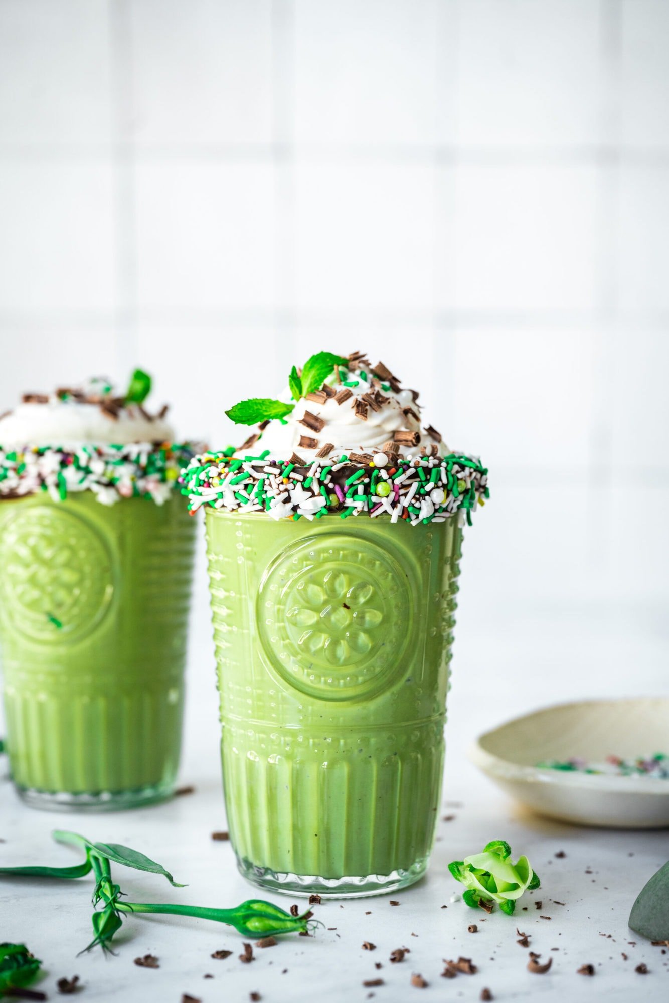 side view of two glasses of vegan shamrock shake with chocolate sprinkle rim and whipped cream on top. 