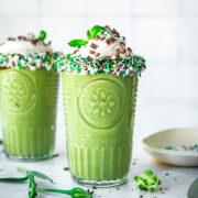 side view of two glasses of vegan shamrock shake with chocolate sprinkle rim and whipped cream on top.