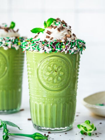 side view of two glasses of vegan shamrock shake with chocolate sprinkle rim and whipped cream on top.