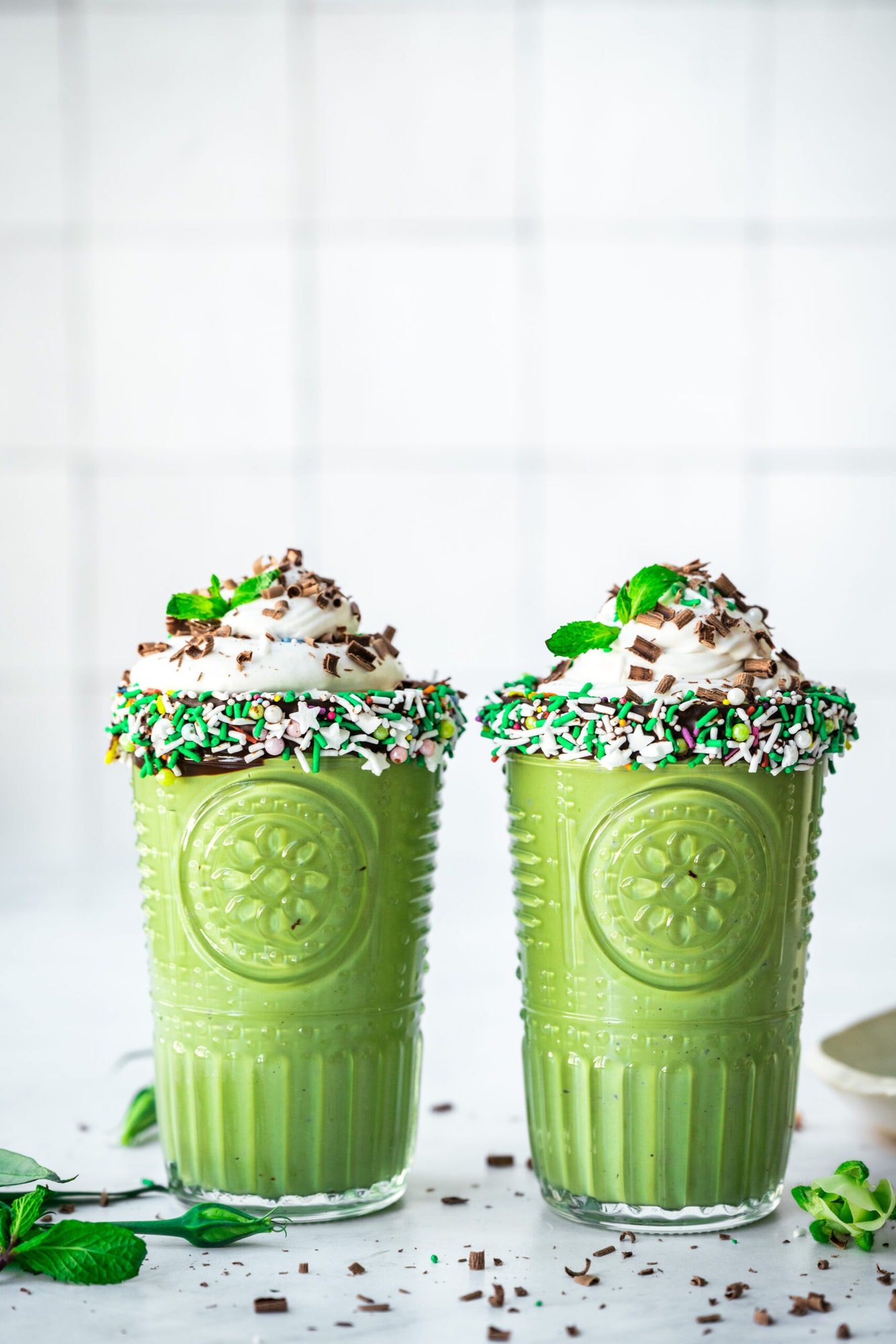 side view of two glasses of vegan shamrock shake with chocolate sprinkle rim and whipped cream on top. 