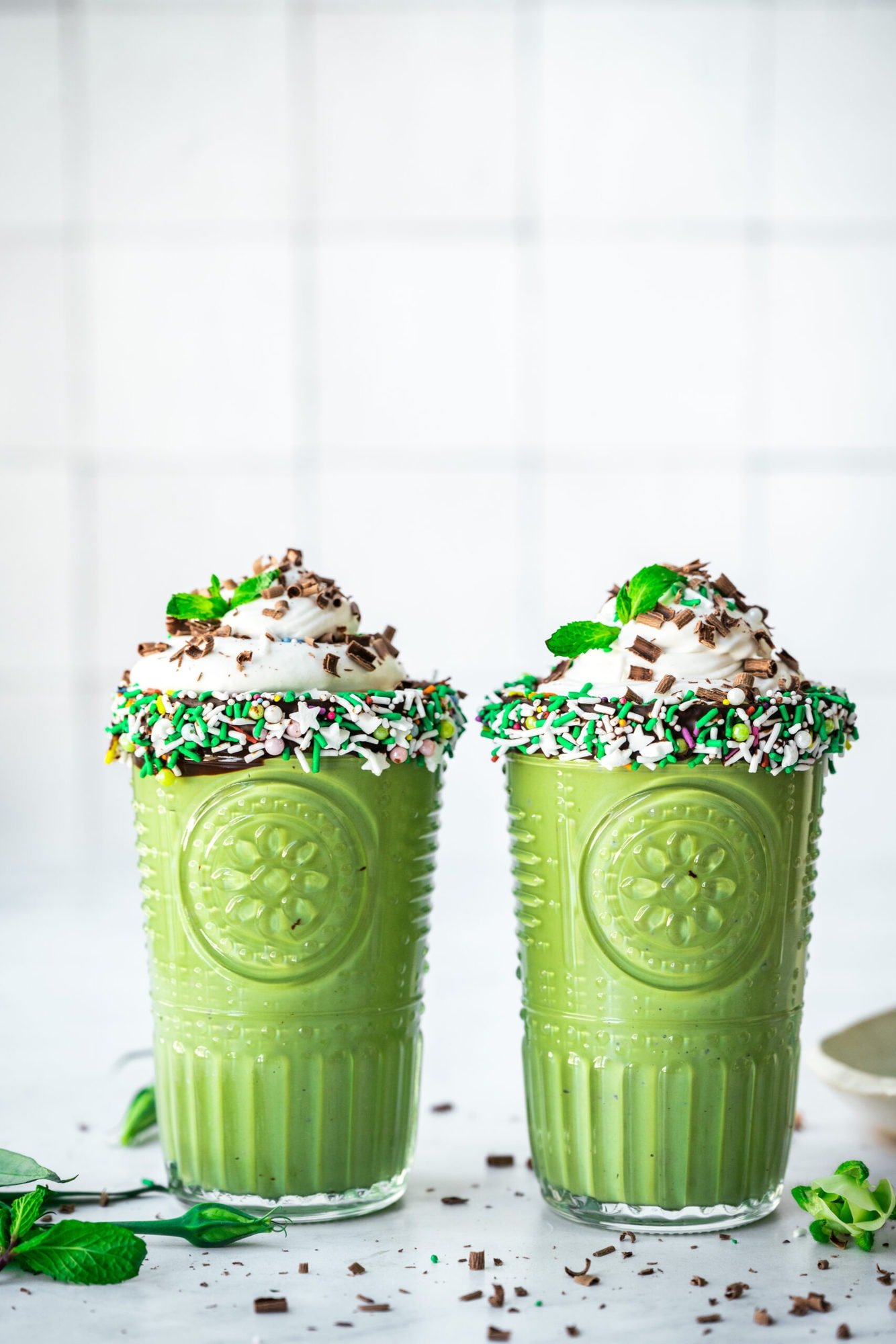 side view of two glasses of vegan shamrock shake with chocolate sprinkle rim and whipped cream on top.