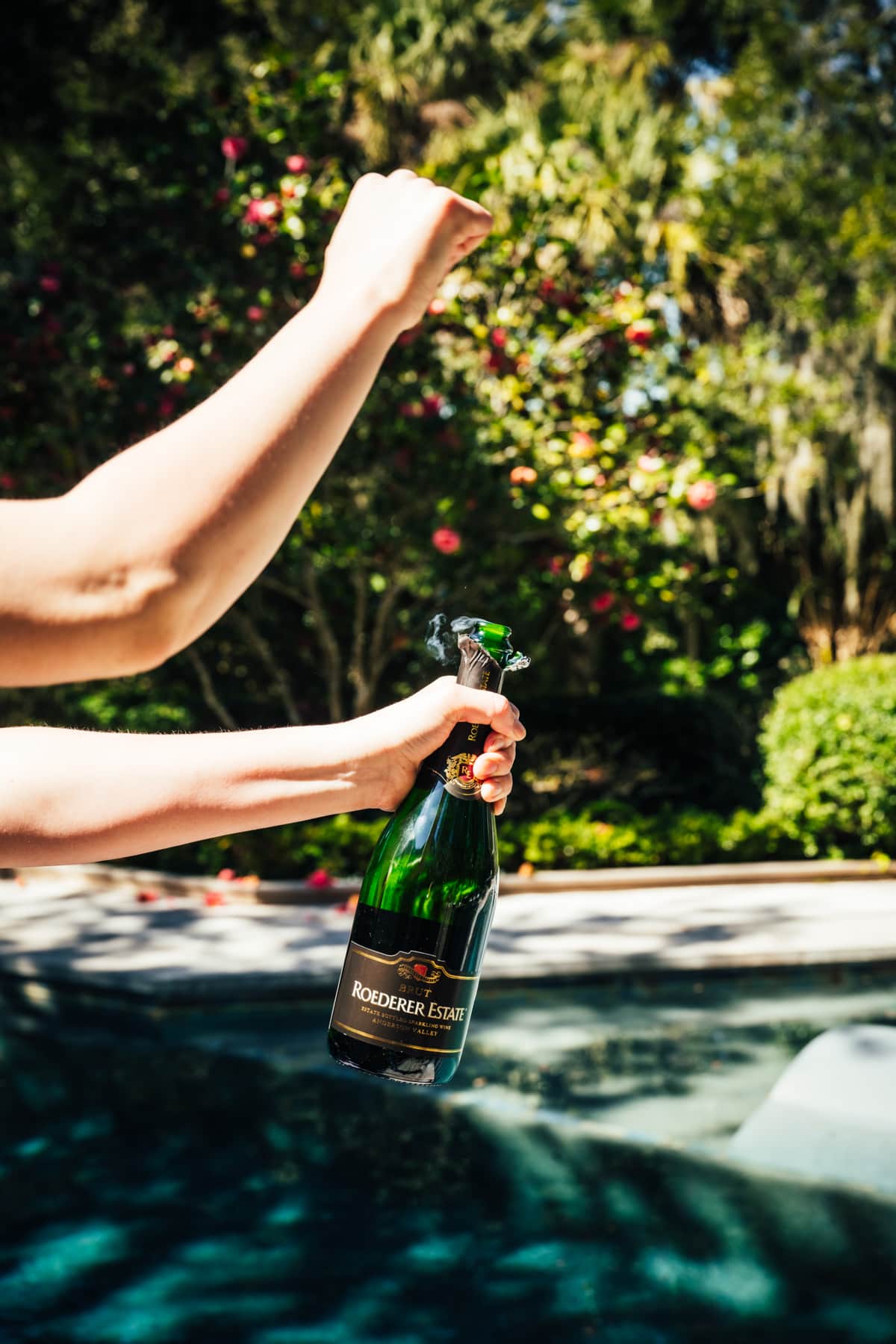 person opening bottle of champagne in front of pool.