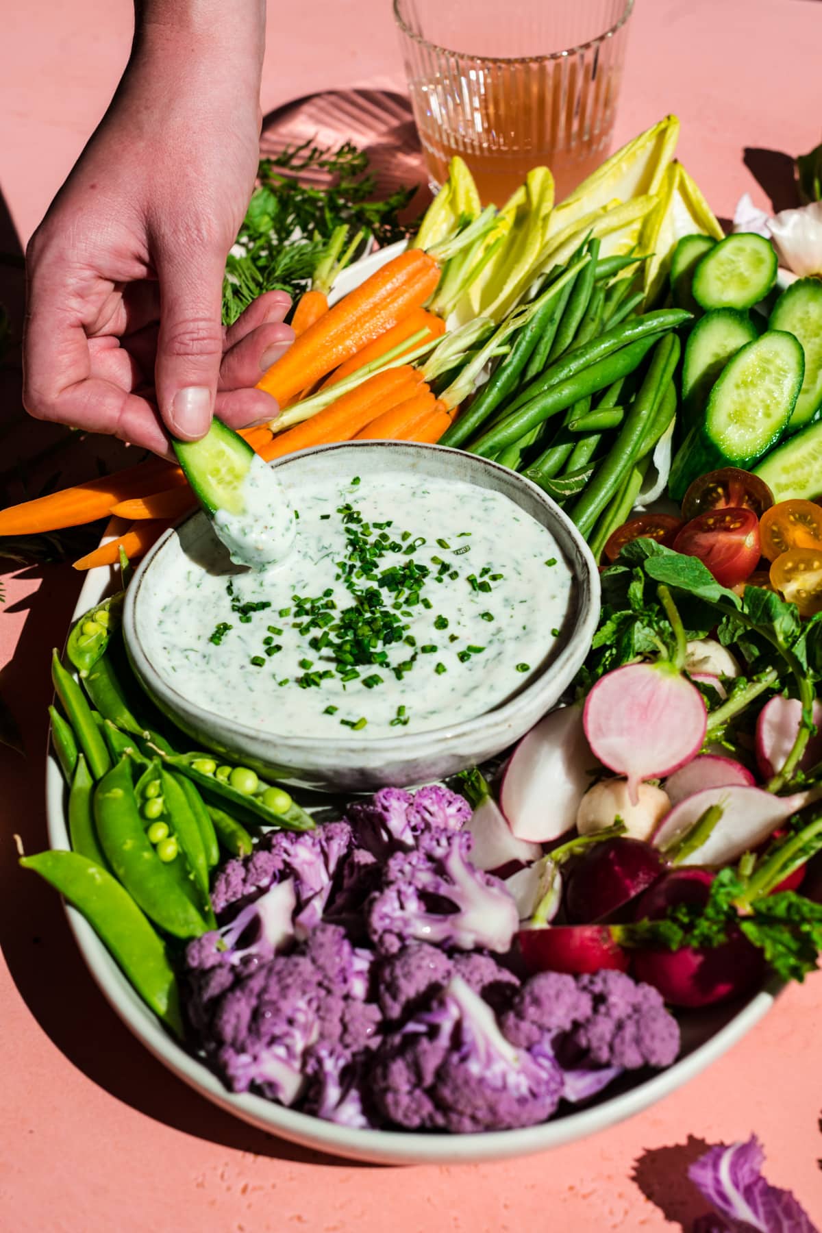 person dipping cucumber slice into vegan ranch dip on a platter with fresh sliced vegetables. 