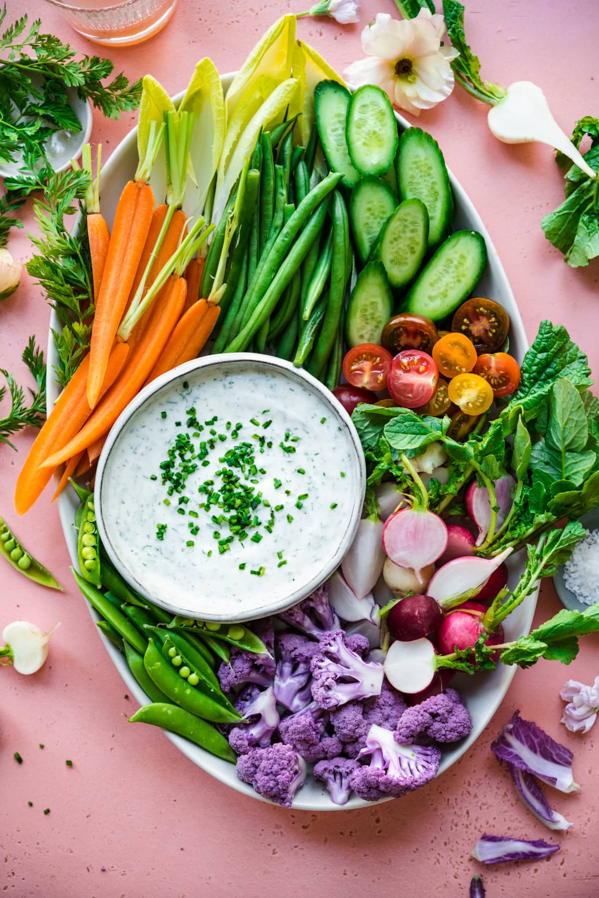 overhead view of vegan ranch dip in bowl with a platter of fresh sliced vegetables on pink backdrop. 