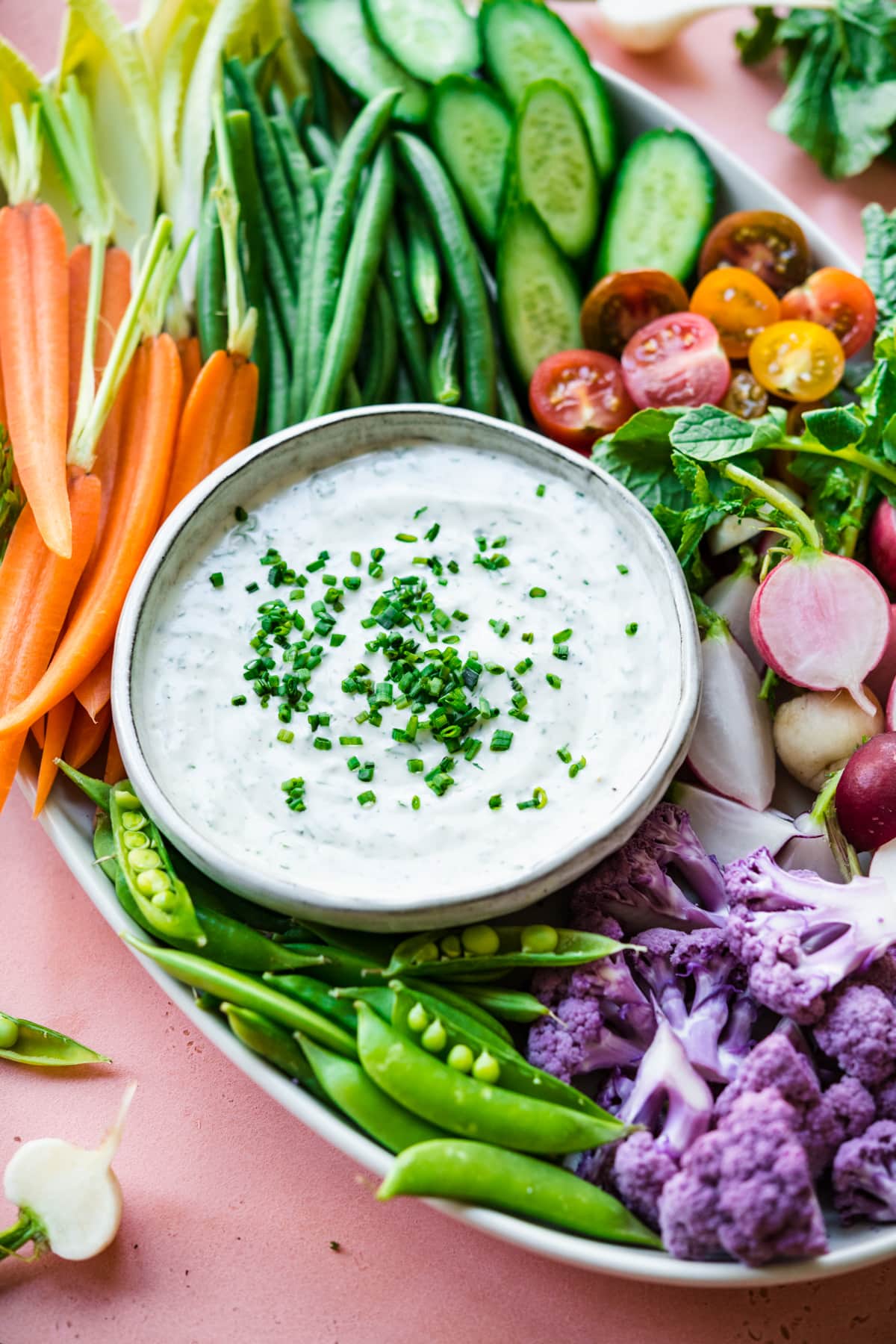 close up view of vegan ranch dip in bowl with a platter of fresh sliced vegetables on pink backdrop. 