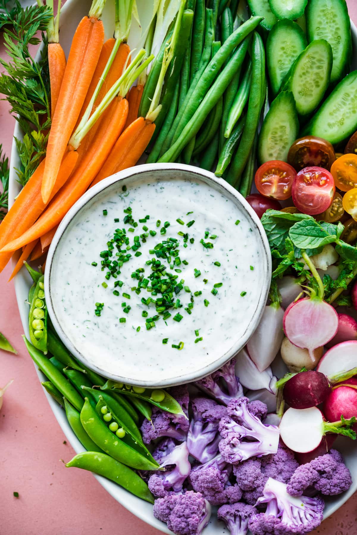 overhead view of vegan ranch dip in bowl with a platter of fresh sliced vegetables on pink backdrop. 