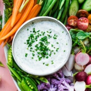 overhead view of vegan ranch dip in bowl with a platter of fresh sliced vegetables on pink backdrop.