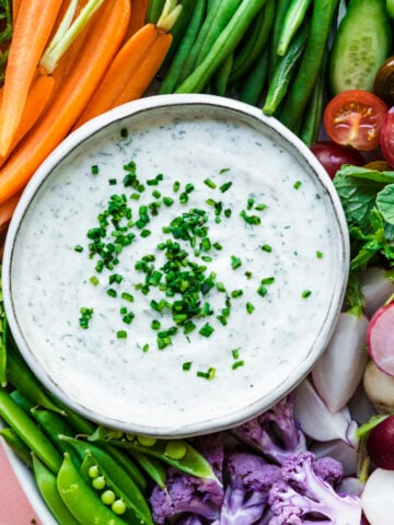 overhead view of vegan ranch dip in bowl with a platter of fresh sliced vegetables on pink backdrop.