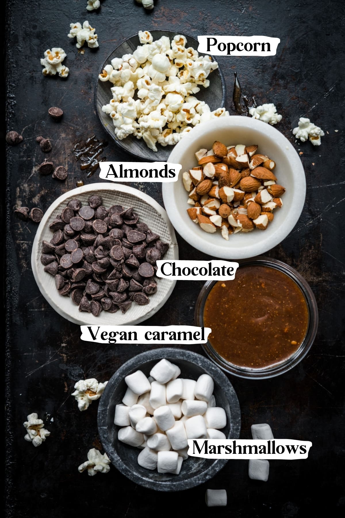 overhead view of popcorn, almonds, chocolate chips, caramel sauce and marshmallows in separate small bowls. 