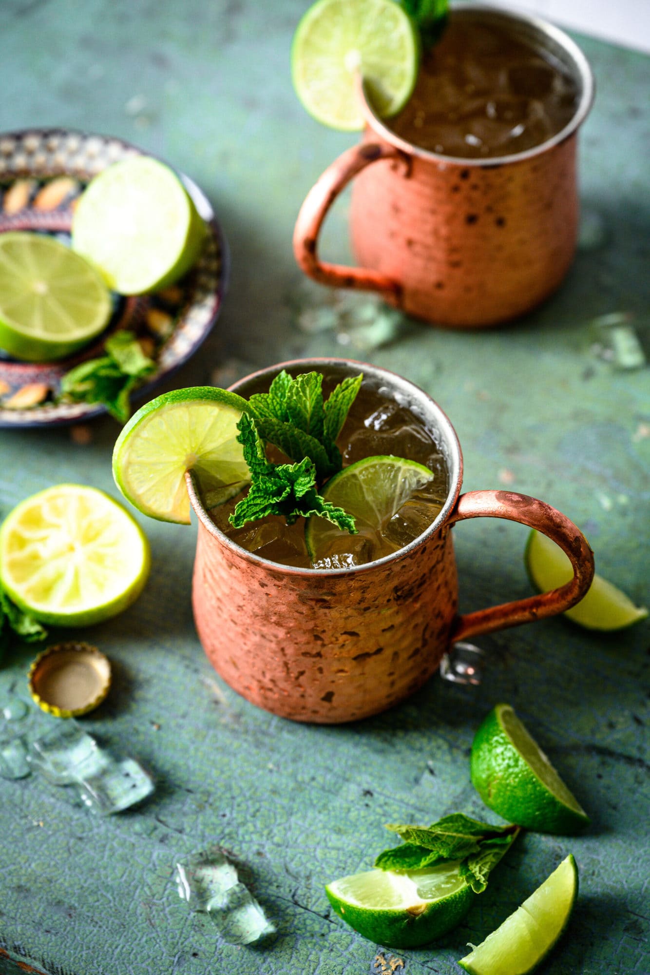 Two tequila mule cocktails in copper mugs with lime and mint garnish on antique blue table. 