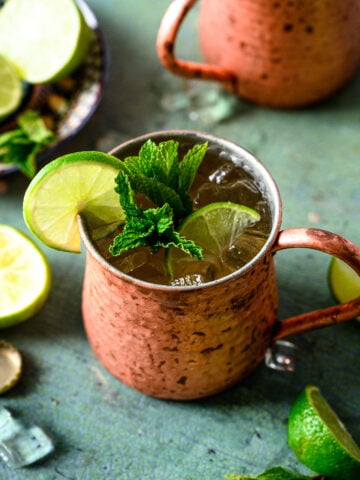 Two tequila mule cocktails in copper mugs with lime and mint garnish on antique blue table.