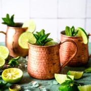 Two tequila mule cocktails in copper mugs with lime and mint garnish on antique blue table.