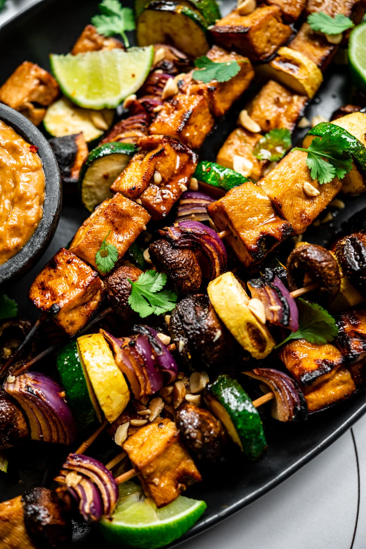 close up view of grilled tofu kebabs with vegetables on a platter with a bowl of peanut sauce. 