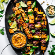 close up view of grilled tofu kebabs with vegetables on a platter with a bowl of peanut sauce.