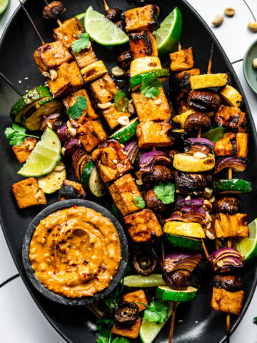 close up view of grilled tofu kebabs with vegetables on a platter with a bowl of peanut sauce.