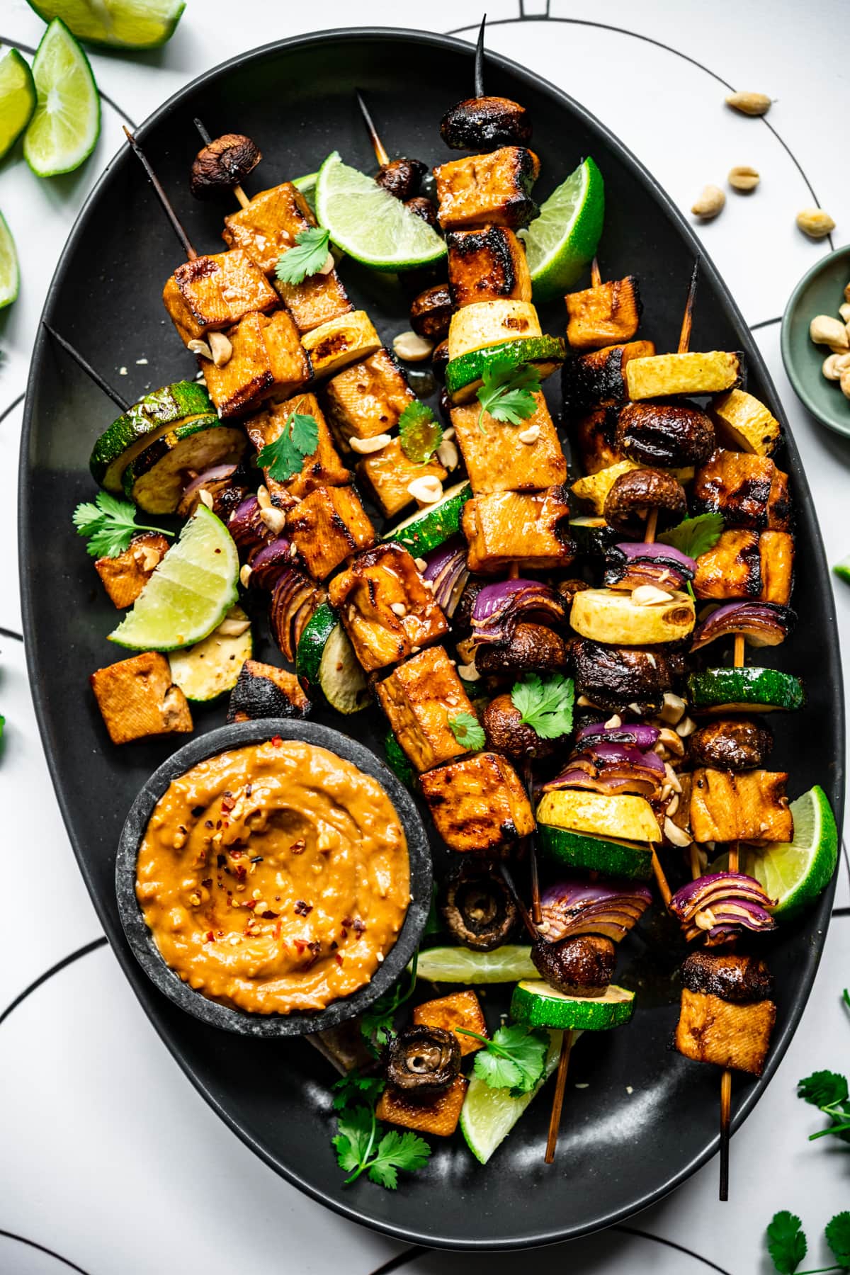 close up view of grilled tofu kebabs with vegetables on a platter with a bowl of peanut sauce. 