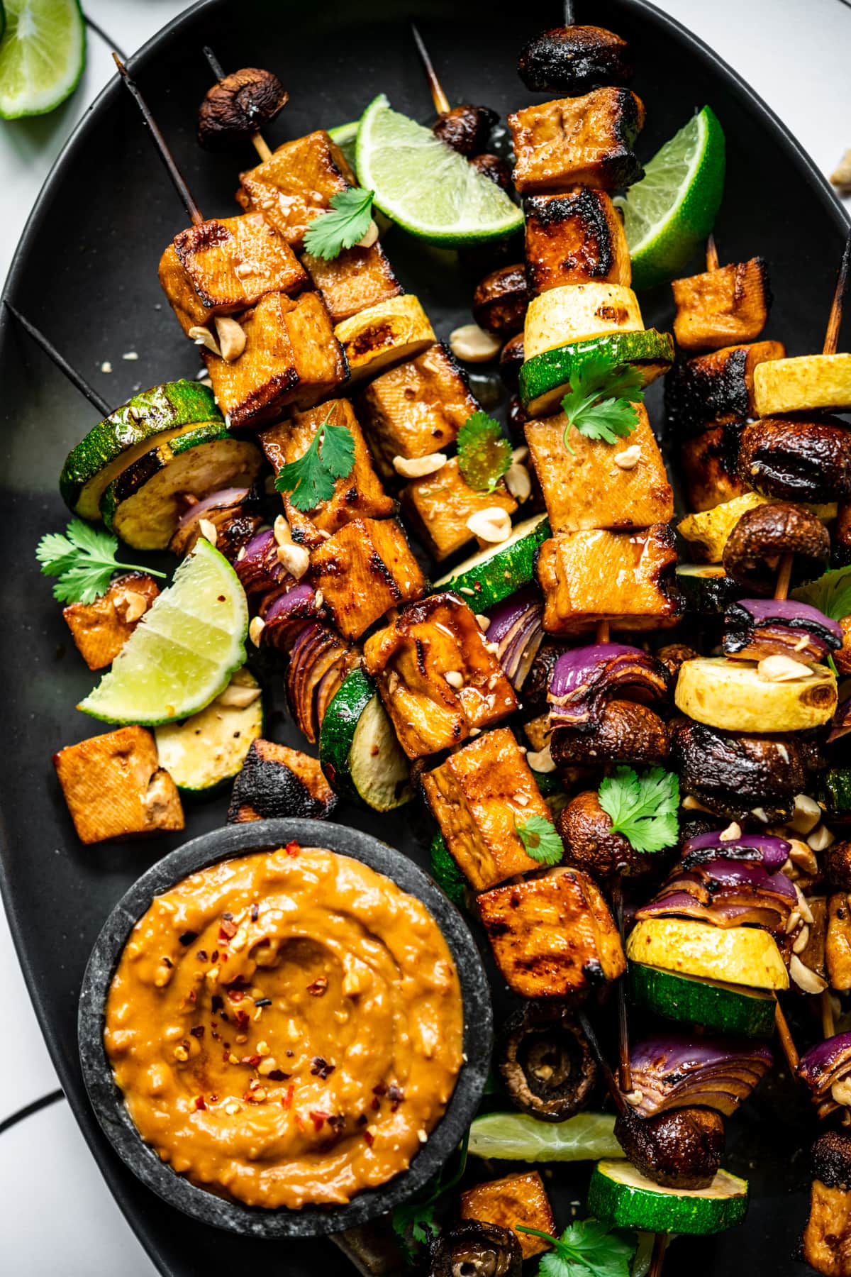 close up view of grilled tofu kebabs with vegetables on a platter with a bowl of peanut sauce. 