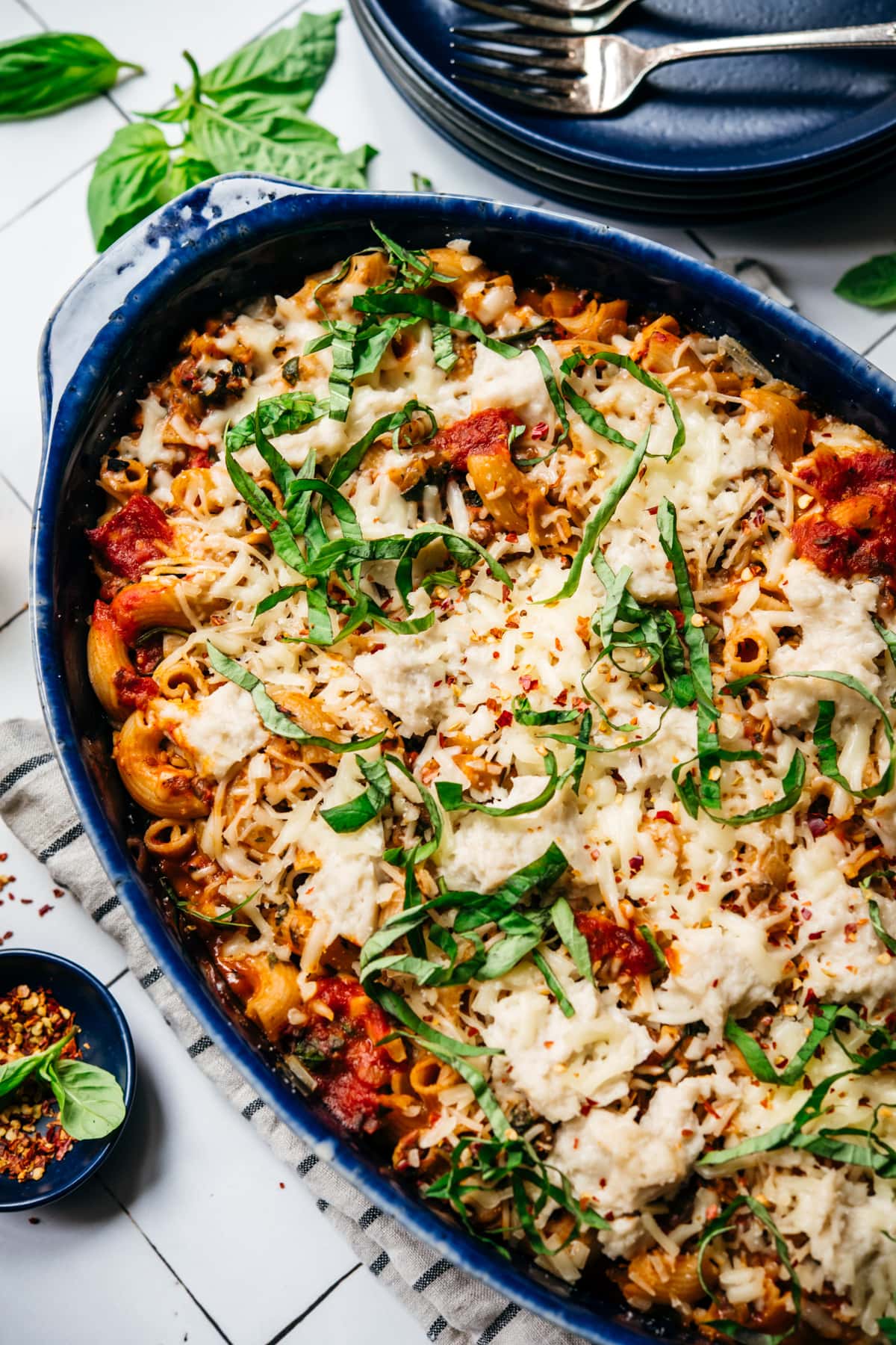 close up view of vegan pasta bake topped with cheese and basil in a blue baking dish. 