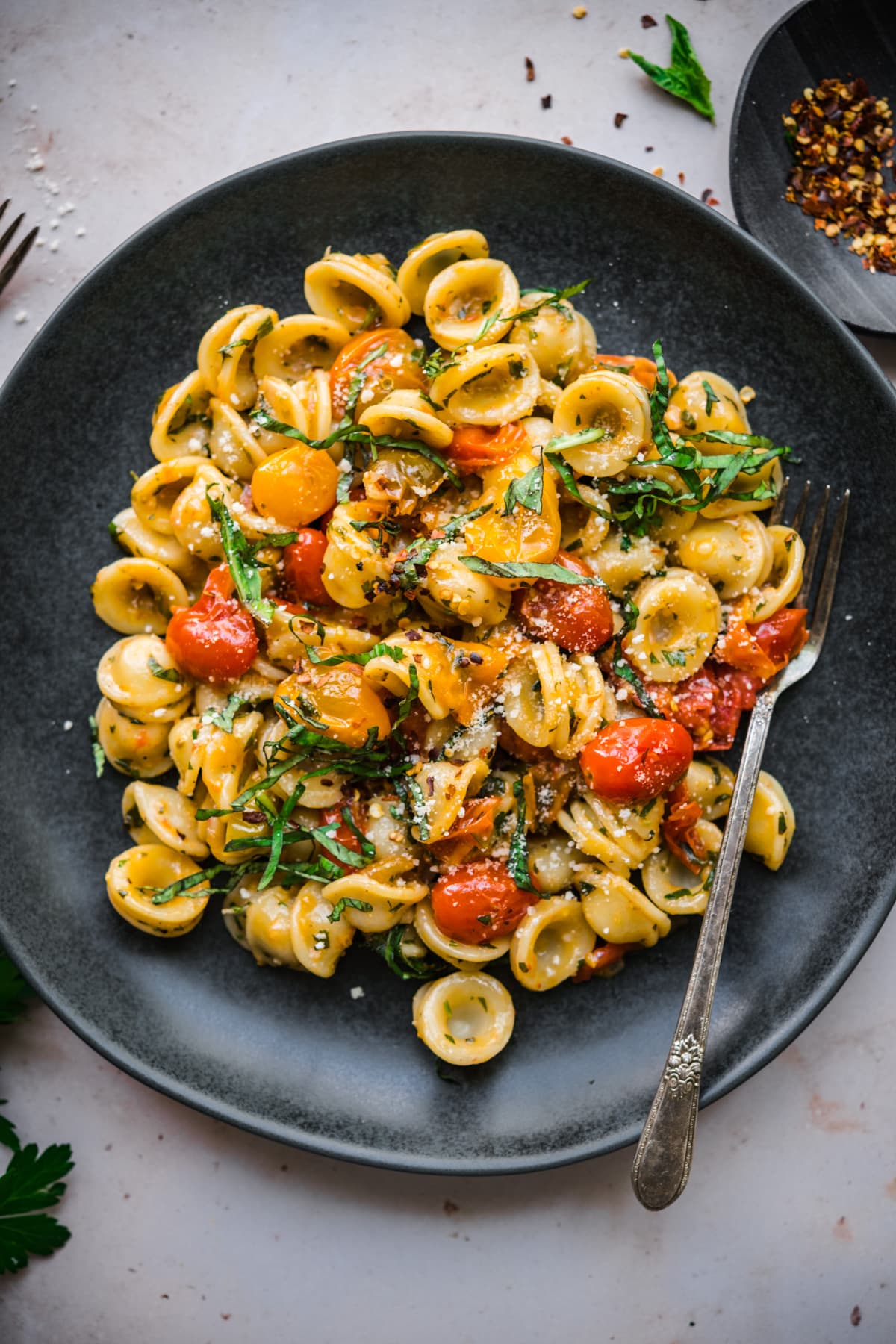 overhead view of burst cherry tomato pasta with fresh basil and red pepper flakes on black plate. 