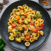 overhead view of burst cherry tomato pasta with fresh basil and red pepper flakes on black plate.