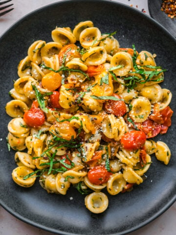 overhead view of burst cherry tomato pasta with fresh basil and red pepper flakes on black plate.