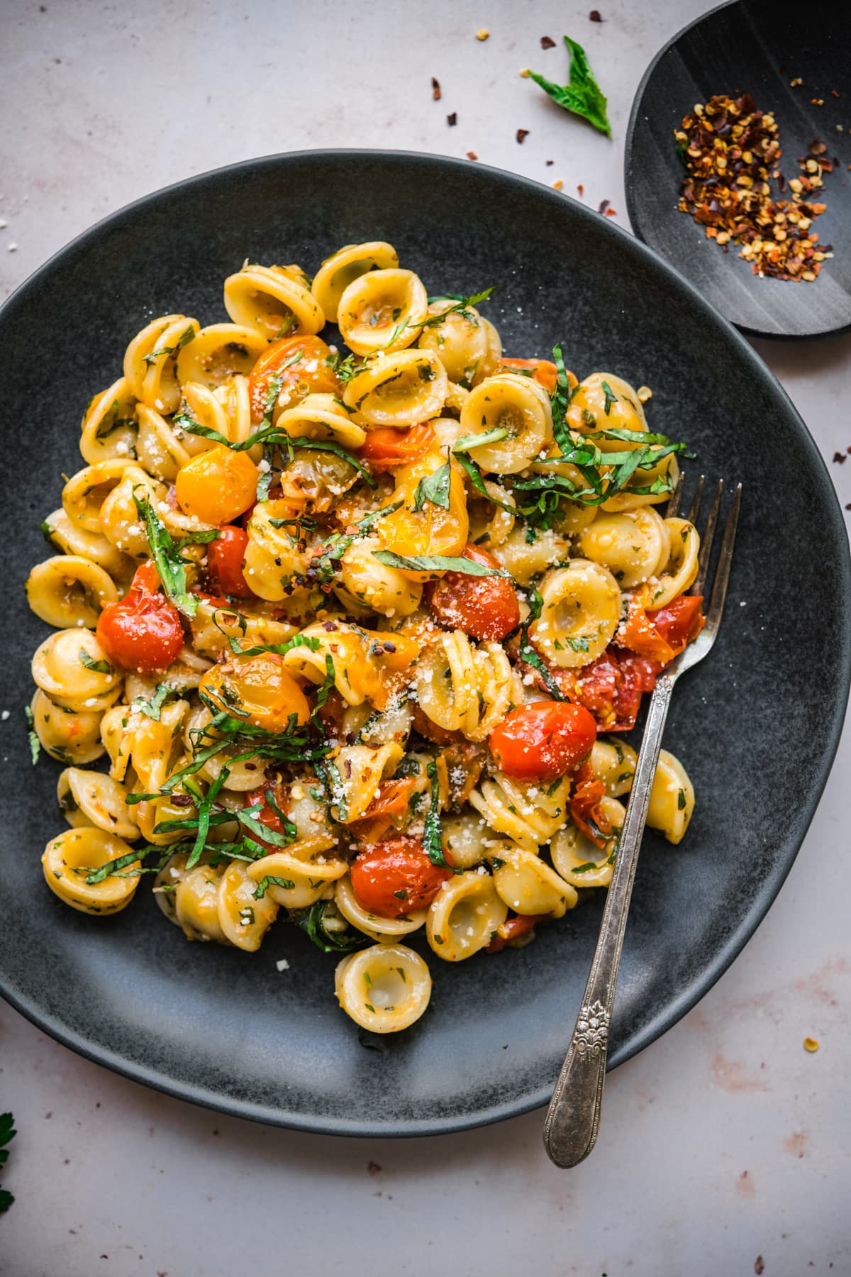 overhead view of burst cherry tomato pasta with fresh basil and red pepper flakes on black plate. 