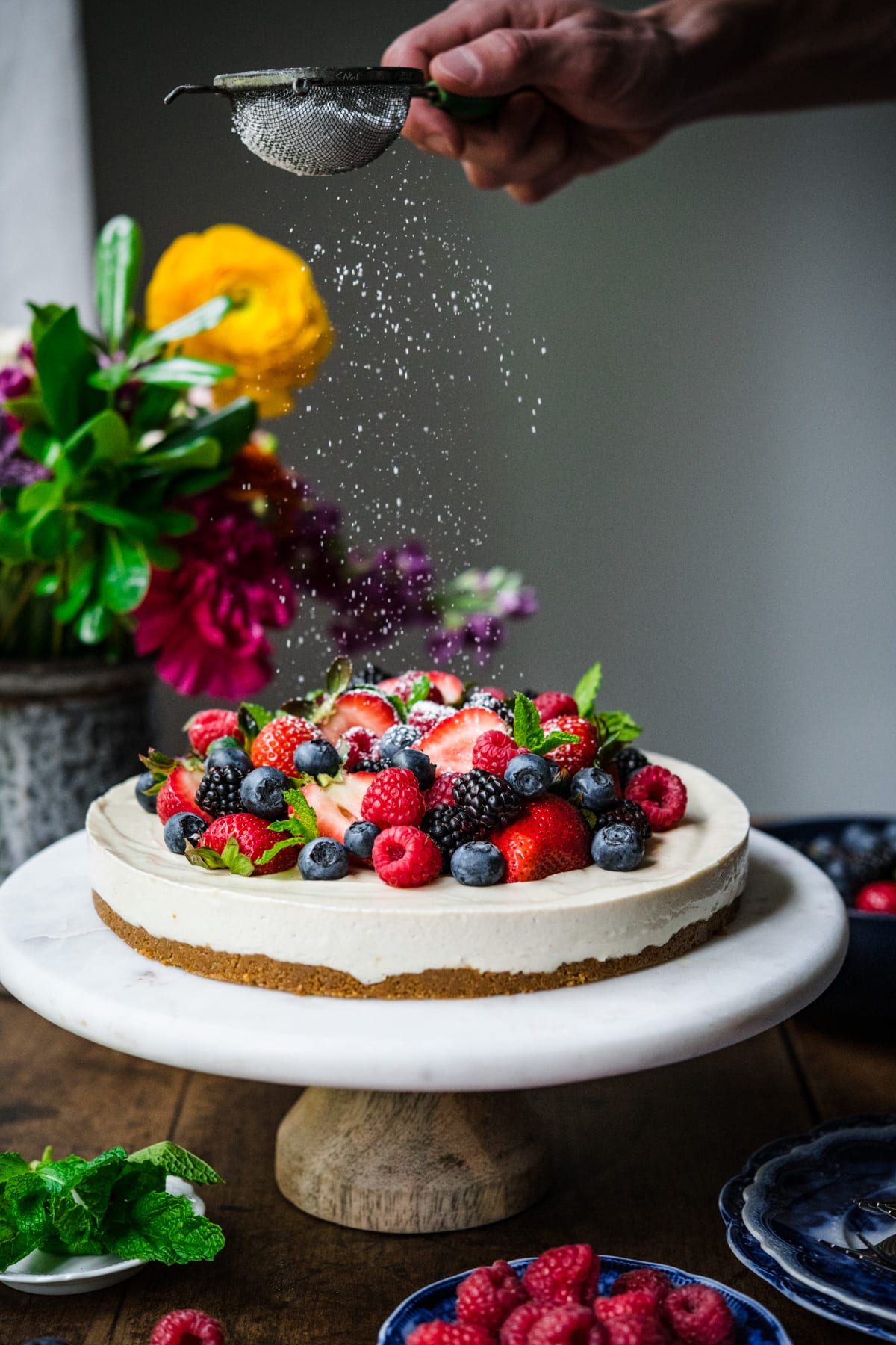 dusting powdered sugar onto no bake vegan cheesecake topped with fresh berries on a marble cake stand. 