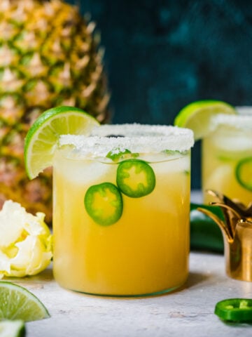 side view of pineapple jalapeño margarita in glass rimmed with salt with a fresh pineapple in background.