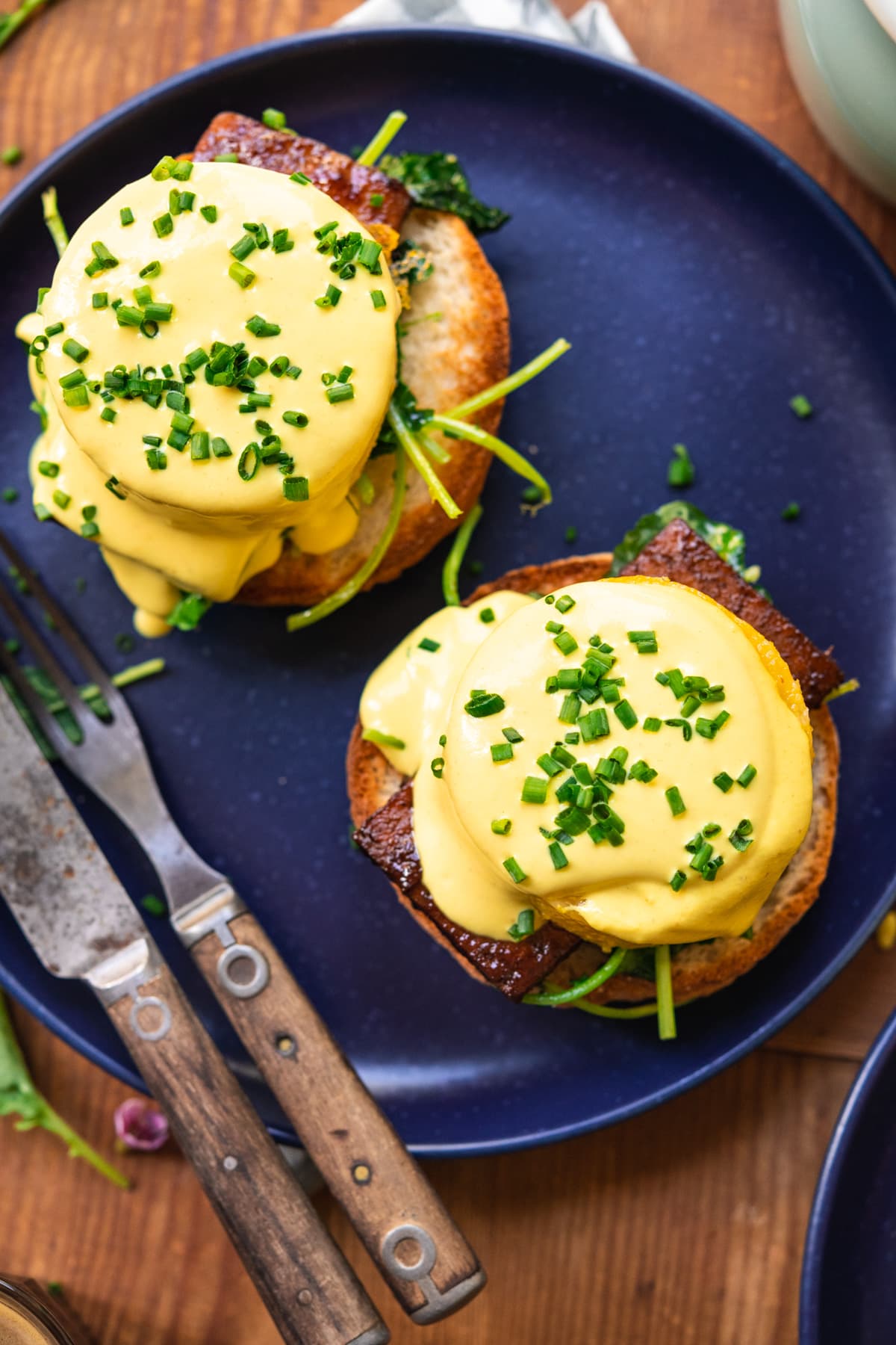 overhead view of vegan eggs benedict on a blue plate with hollandaise sauce and chives. 