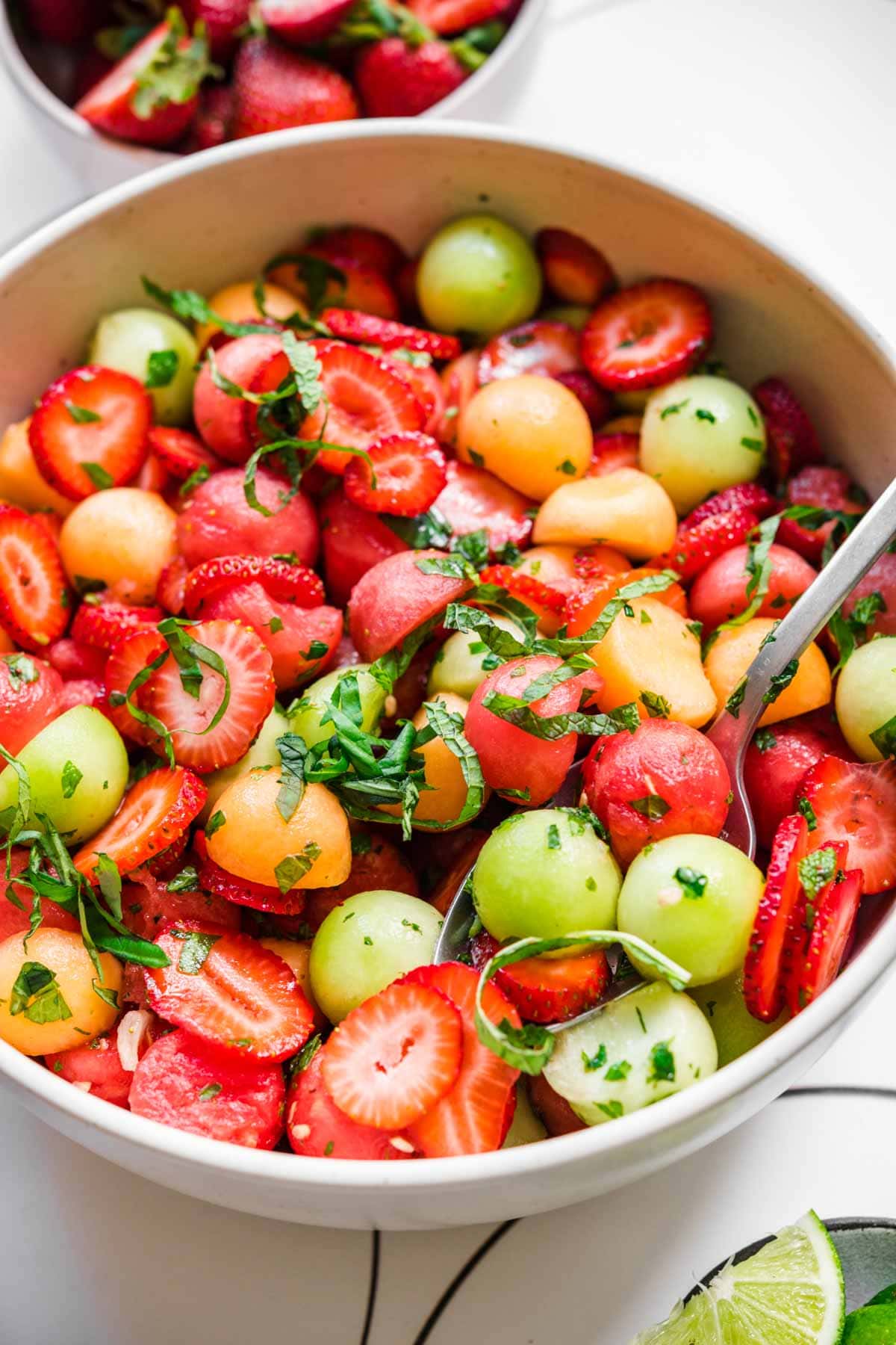 close up side view of melon ball salad with strawberries and mint in white bowl. 