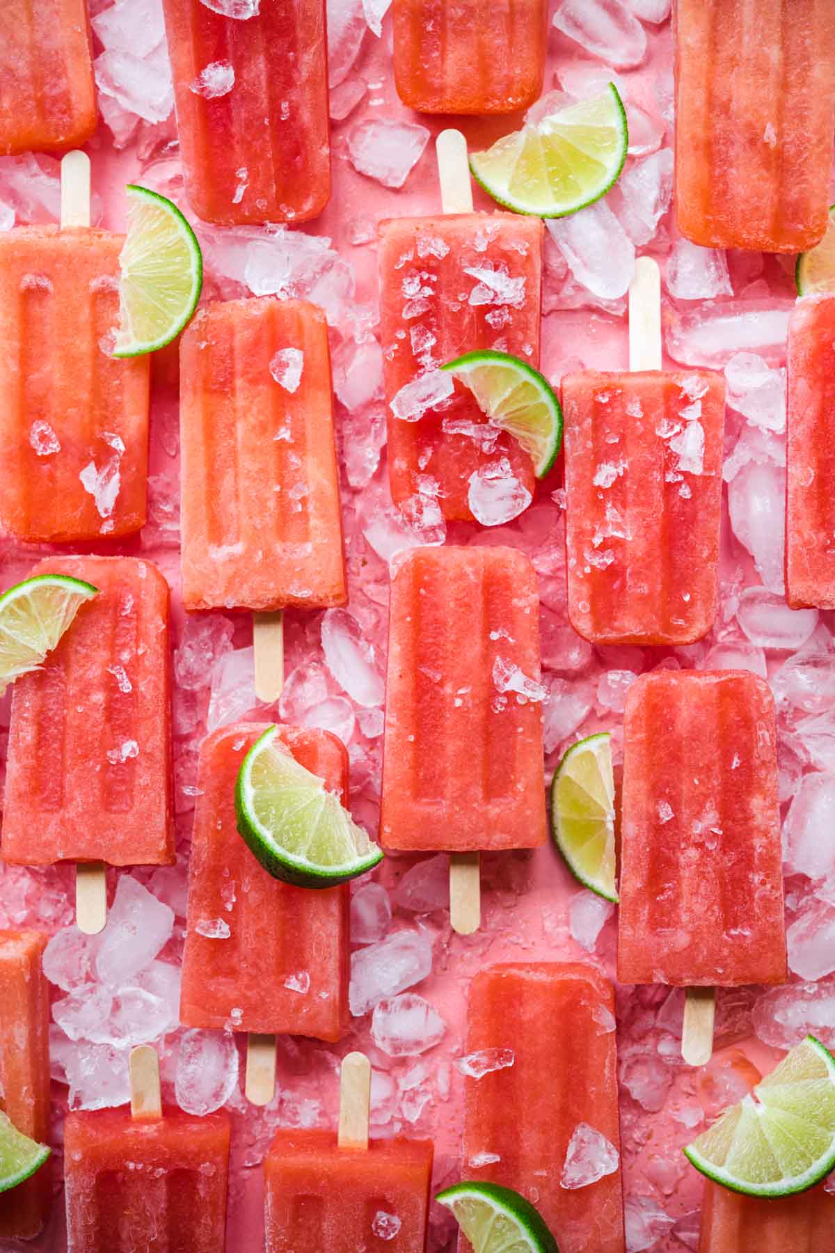 overhead view of watermelon prosecco popsicles on pink backdrop with crushed ice and lime wedges.