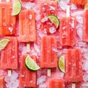 overhead view of watermelon prosecco popsicles on pink backdrop with crushed ice and lime wedges.