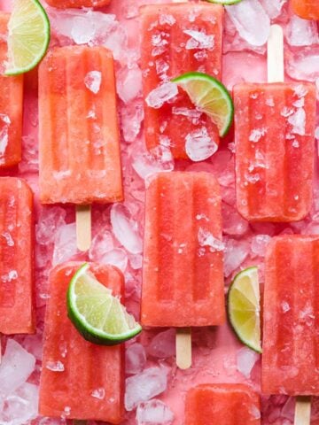 overhead view of watermelon prosecco popsicles on pink backdrop with crushed ice and lime wedges.