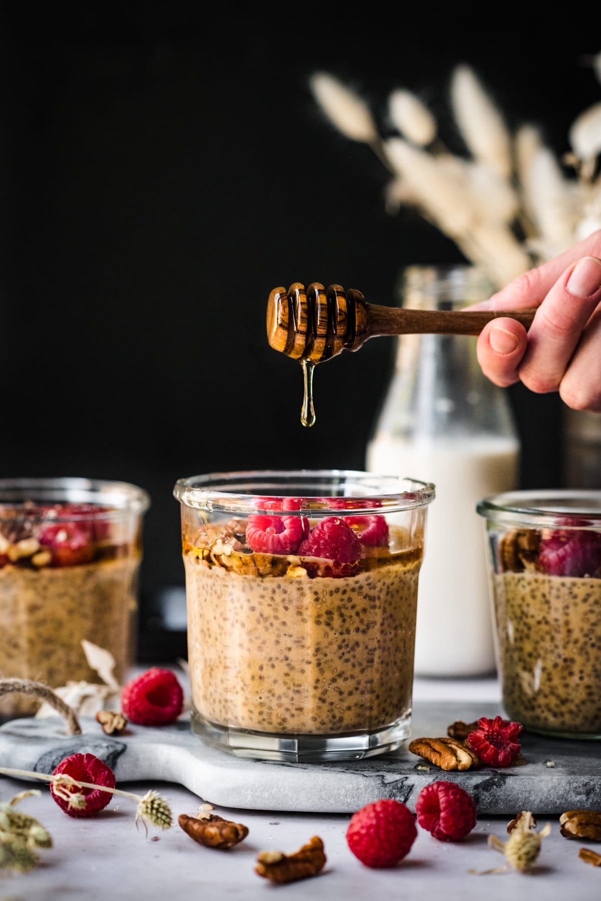 side view of jars of pumpkin chia pudding topped with raspberries and person holding honey drizzler above jar. 