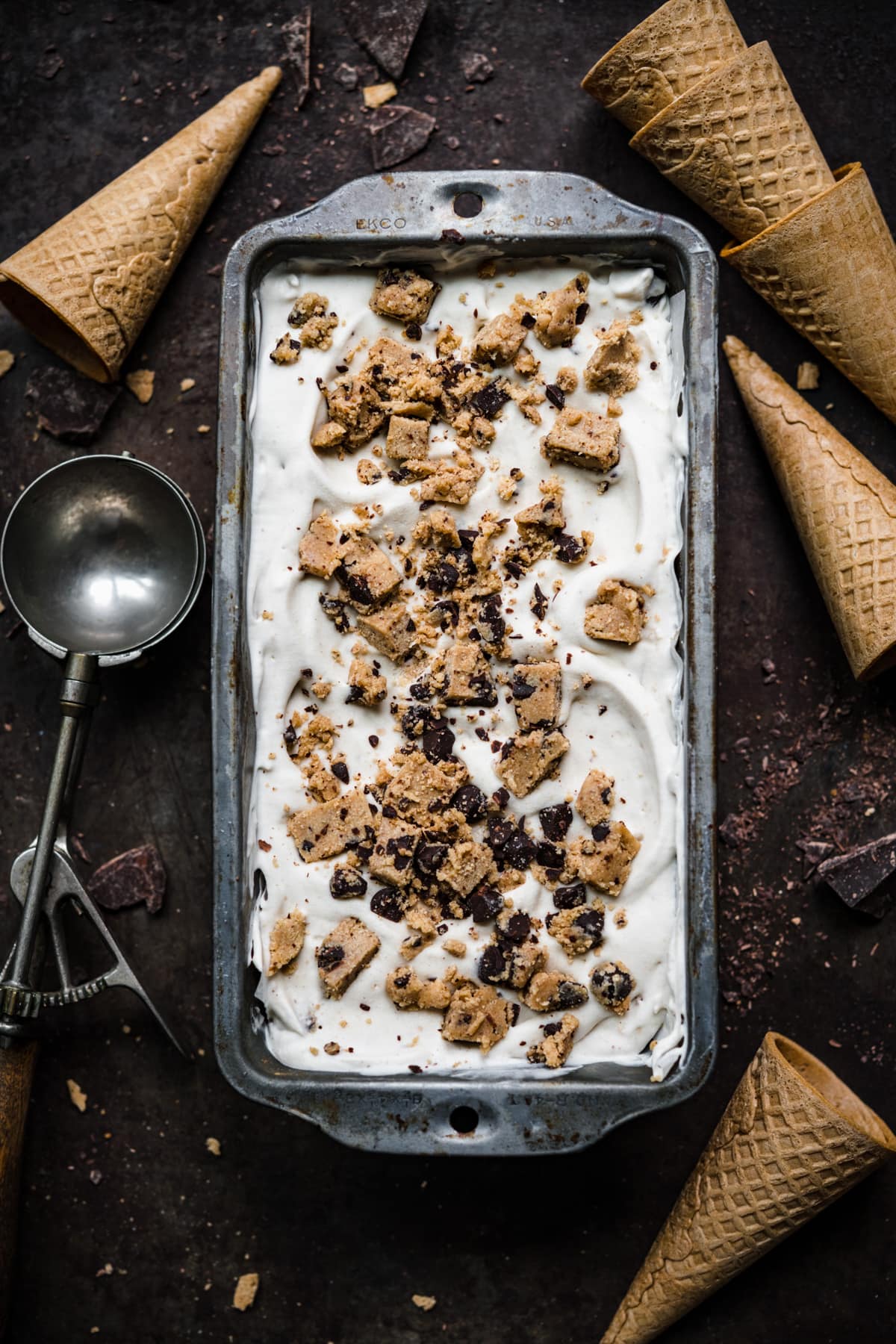 overhead view of vegan cookie dough ice cream in antique tin with ice cream cones around it. 