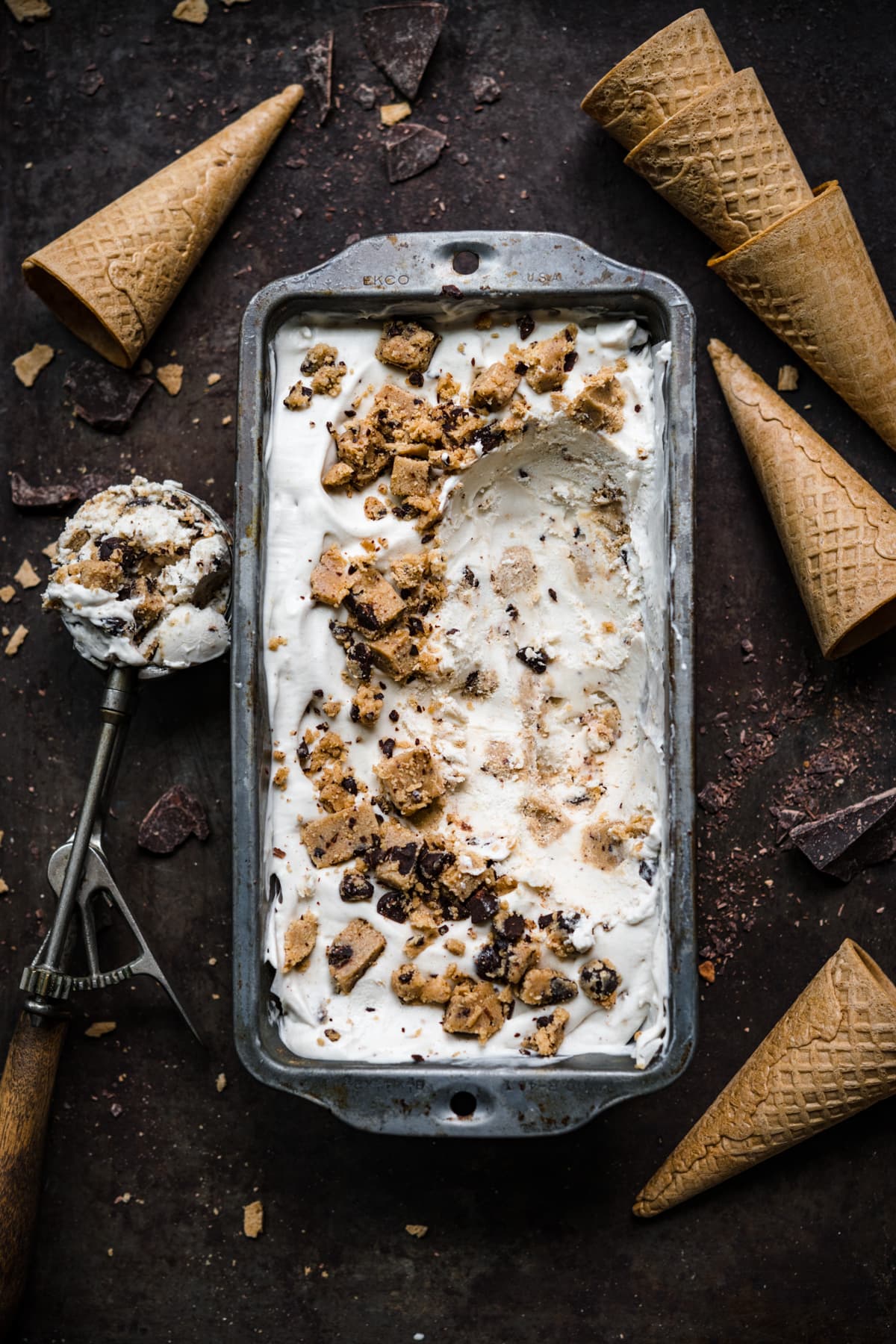 overhead view of vegan cookie dough ice cream in antique tin with ice cream cones around it. 