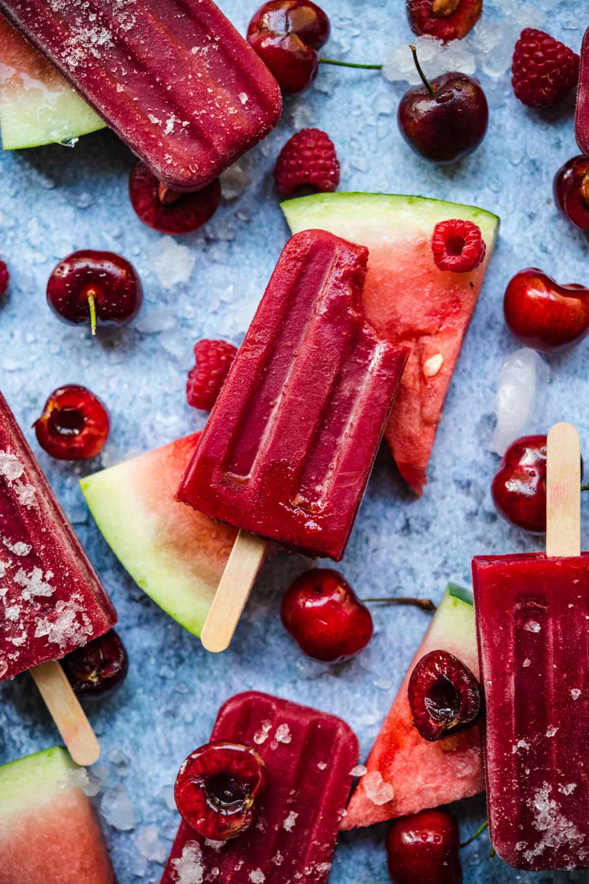 overhead view of watermelon berry popsicles with fresh fruit and ice on blue background.