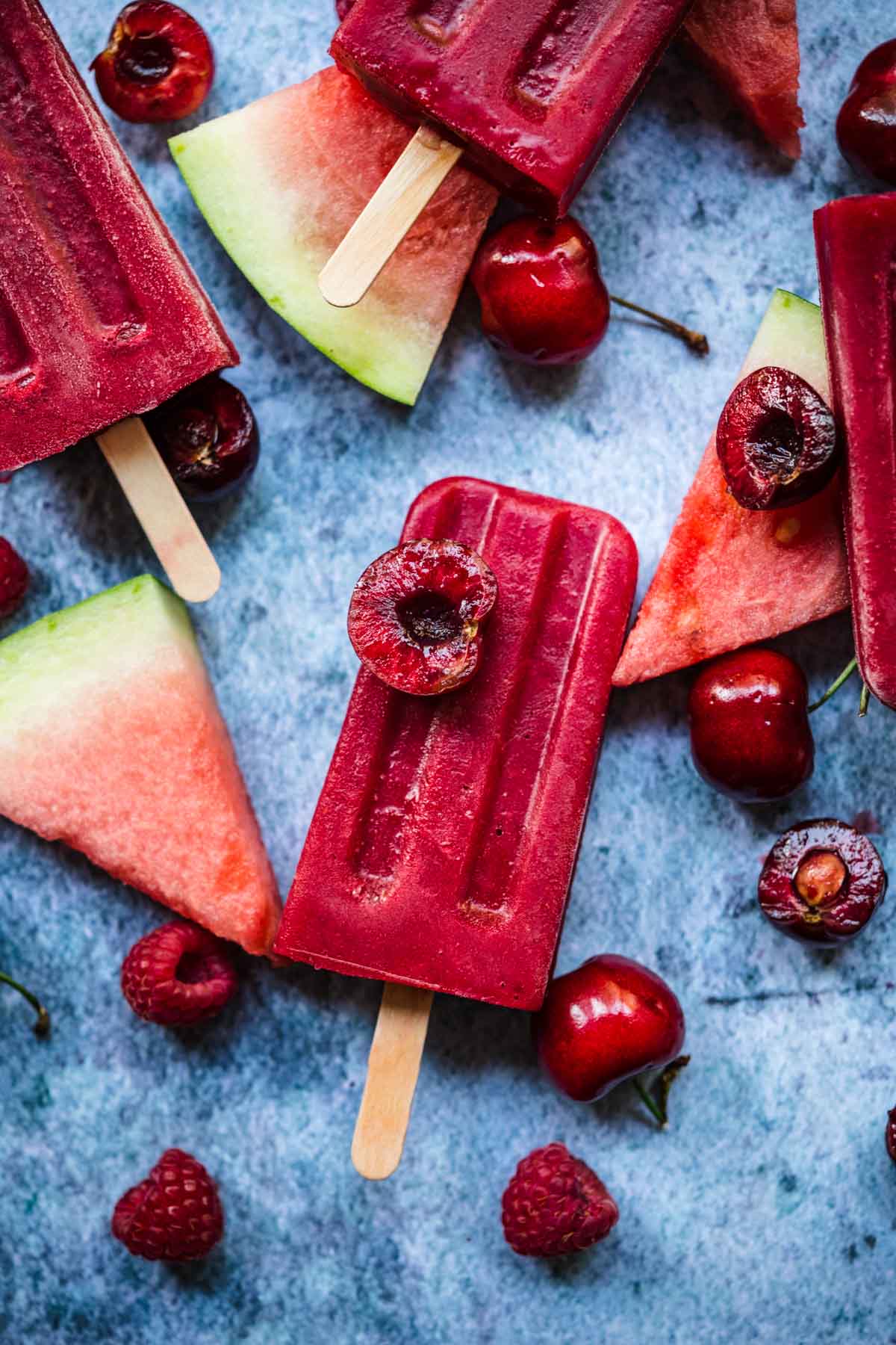 overhead view of watermelon berry popsicles with fresh fruit and ice on blue background.