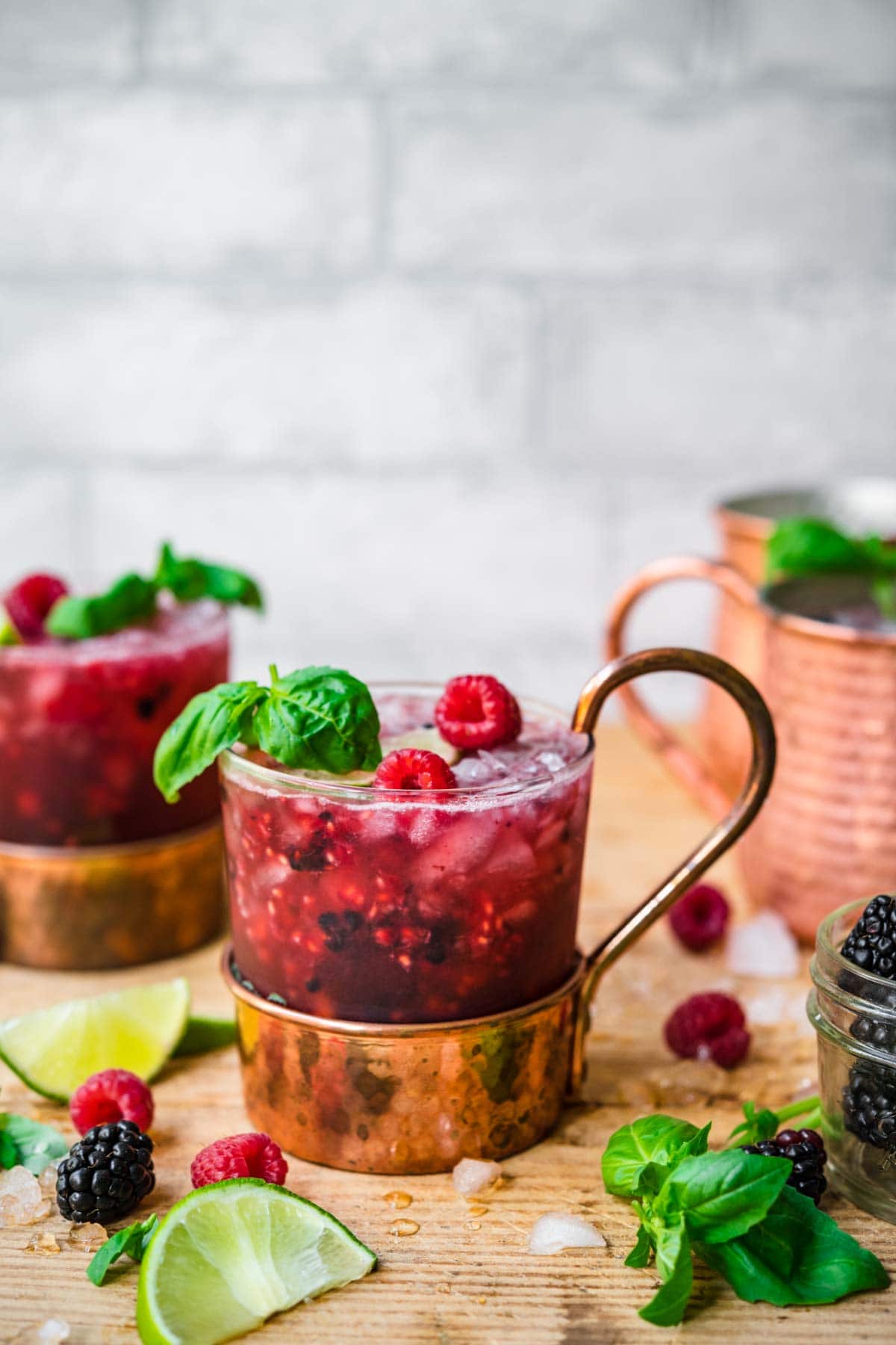 close up side view of berry basil moscow mules garnished with basil and raspberries on wood table. 