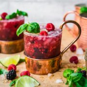close up side view of berry basil moscow mules garnished with basil and raspberries on wood table.