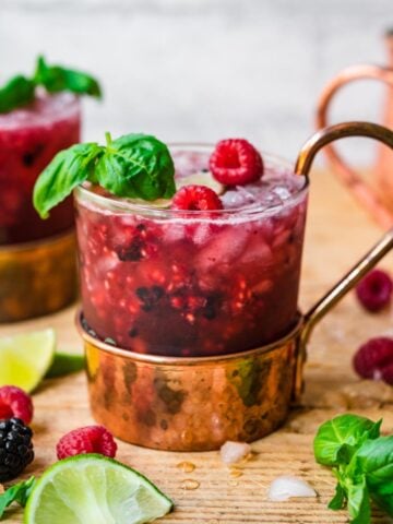 close up side view of berry basil moscow mules garnished with basil and raspberries on wood table.