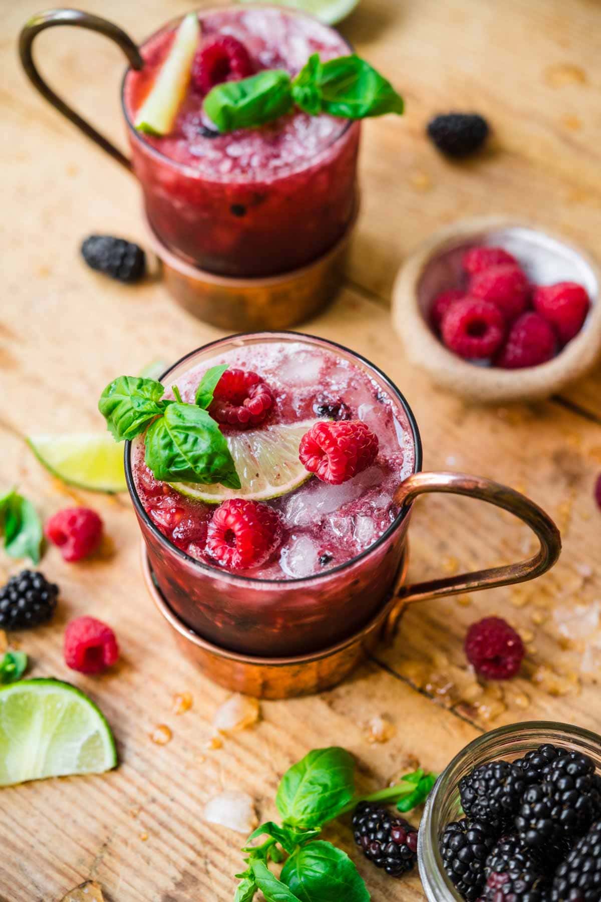 close up side view of berry basil moscow mules garnished with basil and raspberries on wood table. 