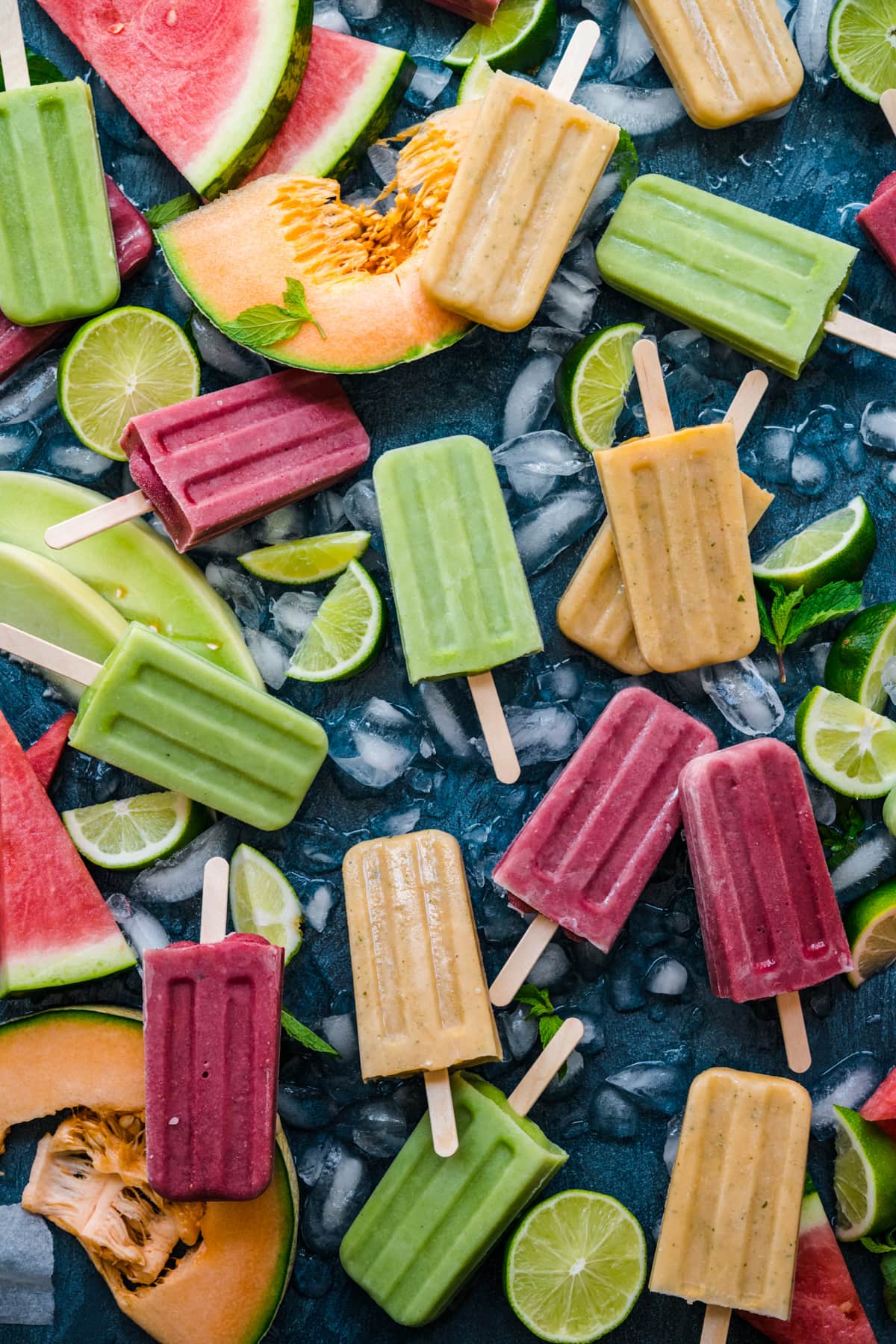 overhead view of watermelon, cantaloupe and honeydew melon yogurt popsicles on blue backdrop with fresh melon. 