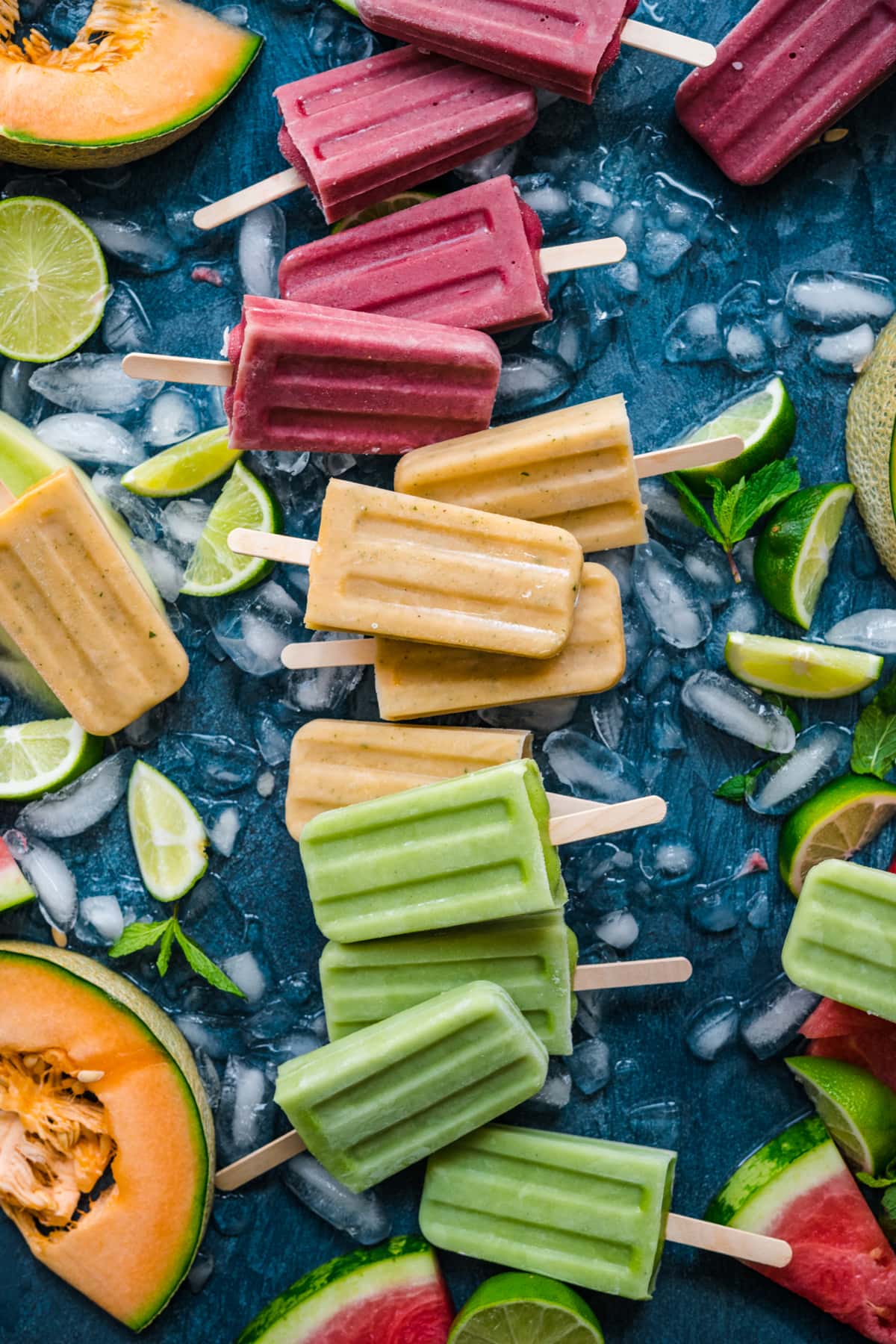overhead view of watermelon, cantaloupe and honeydew melon yogurt popsicles on blue backdrop with fresh melon. 