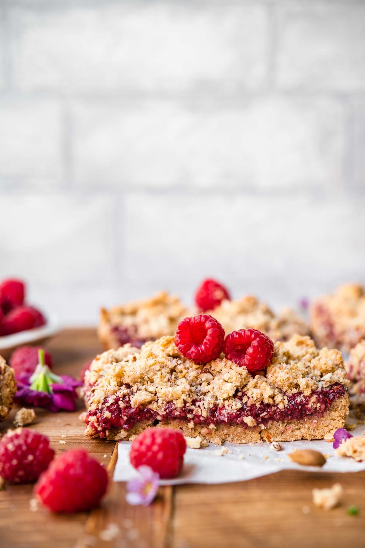 close up side view of raspberry crumble bar topped with fresh raspberries on parchment paper. 