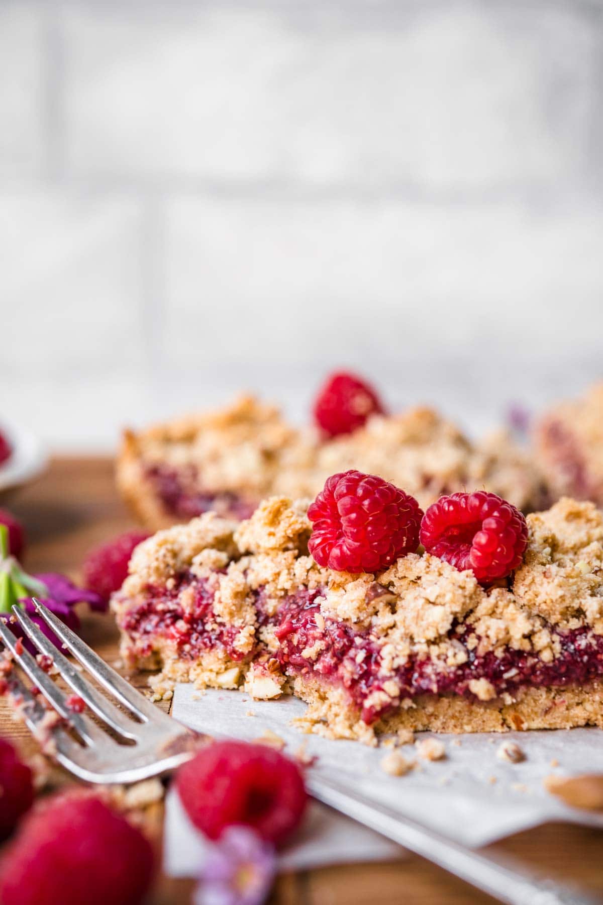 close up side view of raspberry crumble bar topped with fresh raspberries on parchment paper. 