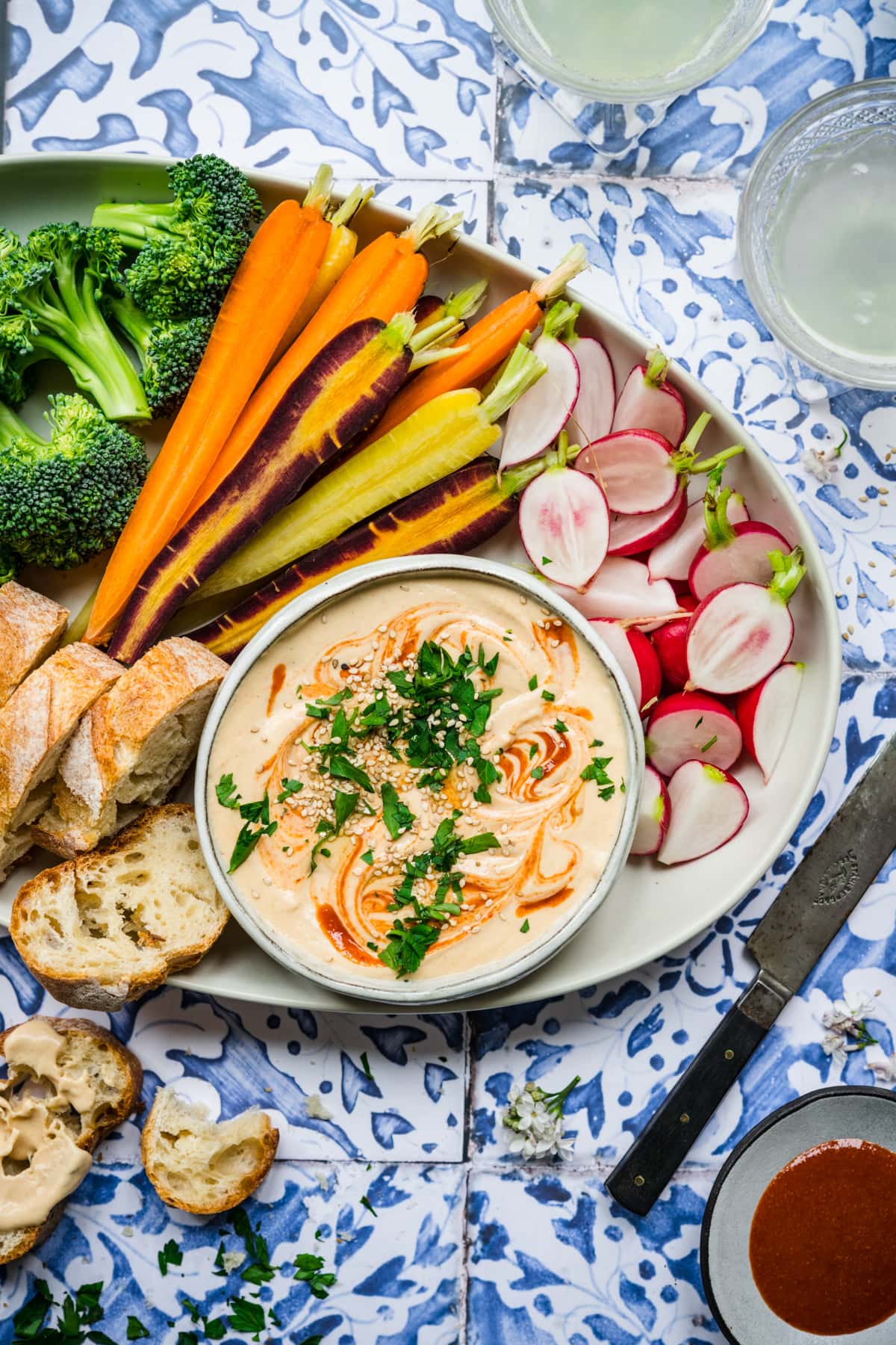 overhead view of sriracha white bean dip on a platter with fresh vegetables on blue tile backdrop.