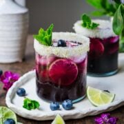 Blueberry margarita in a glass with a salted rim, garnished with mint, blueberries, and lime.