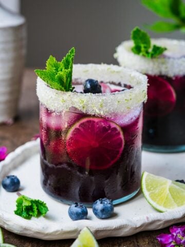 Blueberry margarita in a glass with a salted rim, garnished with mint, blueberries, and lime.